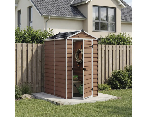 Petit abri de jardin aspect bois, toit à deux pans, porte ouverte avec accessoires, sur socle en béton à côté d'une clôture en bois.