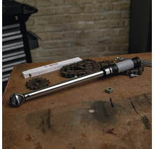 Clé dynamométrique sur un établi en bois avec des outils, une chaîne et un mètre pliant dans un atelier organisé devant un mur en briques.