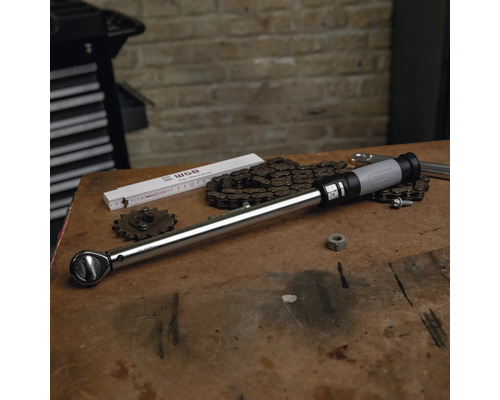 Clé dynamométrique sur un établi en bois avec des outils, une chaîne et un mètre pliant dans un atelier organisé devant un mur en briques.
