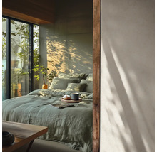 Chambre lumineuse avec linge de lit en lin vert, grandes fenêtres et ombres de plantes sur le mur. Style minimaliste.