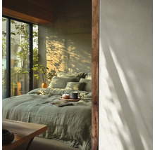 Chambre lumineuse avec lit en lin vert sauge, touches de bois et grande fenêtre donnant sur de la verdure sous la lumière du soleil.