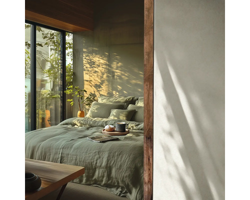 Chambre lumineuse avec lit en lin vert sauge, touches de bois et grande fenêtre donnant sur de la verdure sous la lumière du soleil.