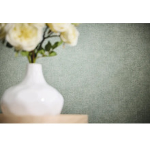 Papier peint intissé uni avec une structure de lin fine dans une nuance de vert clair derrière un vase blanc avec des fleurs.