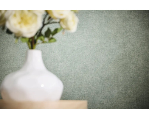 Papier peint intissé uni avec une structure de lin fine dans une nuance de vert clair derrière un vase blanc avec des fleurs.
