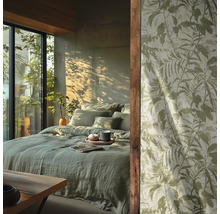 Chambre avec literie en lin vert clair et papier peint à motif de feuilles botaniques devant une fenêtre ensoleillée.