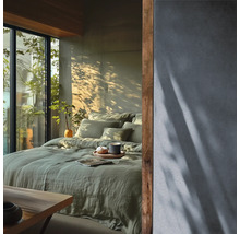 Chambre lumineuse avec parure de lit en lin vert, mur texture et accents en bois a cote d une fenetre avec vue sur la verdure.