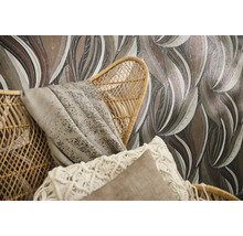 Papier peint intissé à motif de grandes feuilles dans des tons de terre derrière un fauteuil en rotin tressé avec coussins.