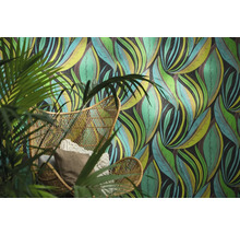 Papier peint intissé avec motif de feuilles abstrait derrière un fauteuil en rotin avec des coussins et une plante verte.