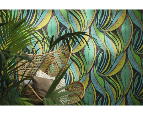Papier peint intissé avec motif de feuilles abstrait derrière un fauteuil en rotin avec des coussins et une plante verte.