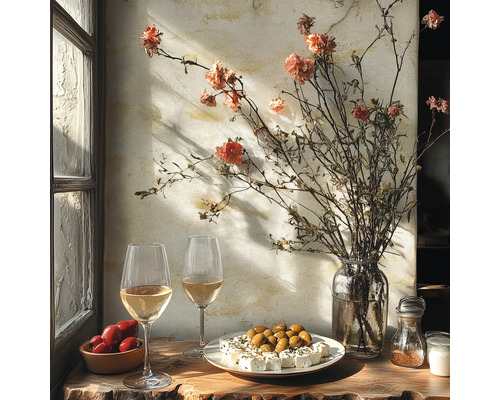 Table rustique près d'une fenêtre avec vin blanc, olives, féta, tomates et un vase de fleurs séchées devant un mur texturé.