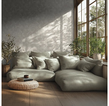 Salon moderne avec canapé modulaire, nombreux coussins, papier peint à motifs, parquet, pouf tressé et plantes près d'une fenêtre ensoleillée.