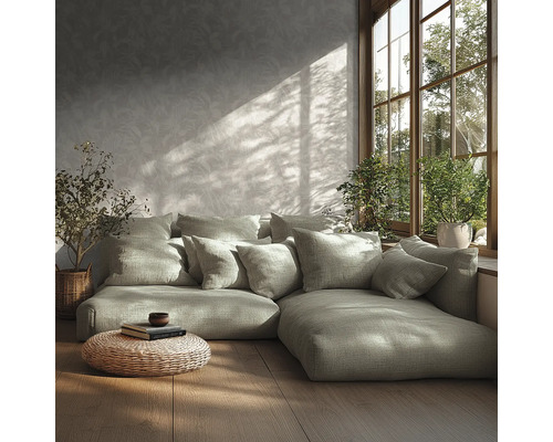 Salon moderne avec canapé modulaire, nombreux coussins, papier peint à motifs, parquet, pouf tressé et plantes près d'une fenêtre ensoleillée.