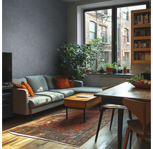 Salon moderne avec mur gris aspect béton, canapé gris, table en bois, tapis oriental et grandes plantes d'intérieur devant une fenêtre.