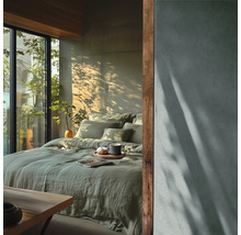 Chambre à coucher moderne avec parure de lit en lin vert, grande baie vitrée donnant sur des plantes et mur à effet béton avec jeux d'ombre.