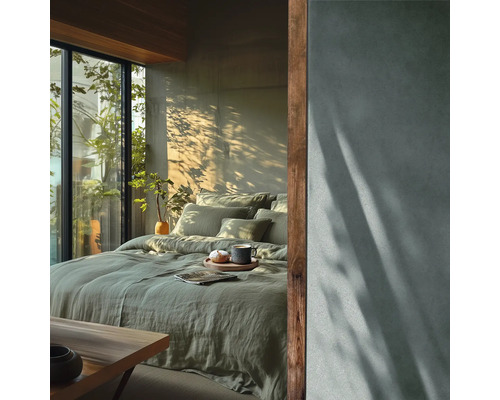 Chambre à coucher moderne avec parure de lit en lin vert, grande baie vitrée donnant sur des plantes et mur à effet béton avec jeux d'ombre.