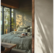 Chambre moderne avec lit en lin vert, mur aspect béton avec ombres d'arbres et grande fenêtre avec plantes.