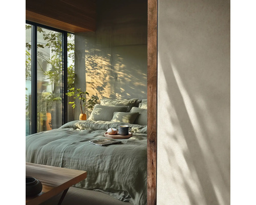 Chambre moderne avec lit en lin vert, mur aspect béton avec ombres d'arbres et grande fenêtre avec plantes.