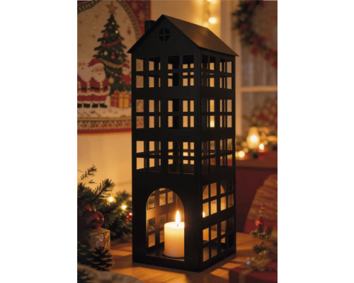 Lanterne en métal noir en forme de maison avec bougie blanche allumée dans un décor de Noël festif avec branches de sapin et lumières chaudes.