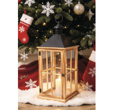 Lanterne en bois avec vitres et toit en métal noir devant un sapin de Noël décoré avec une bougie allumée à l'intérieur.