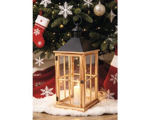 Lanterne en bois avec vitres et toit en métal noir devant un sapin de Noël décoré avec une bougie allumée à l'intérieur.