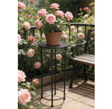 Porte-plante rond et haut en metal noir avec ornements portant un pot en terre cuite avec des roses roses dans un jardin lumineux pres du banc.
