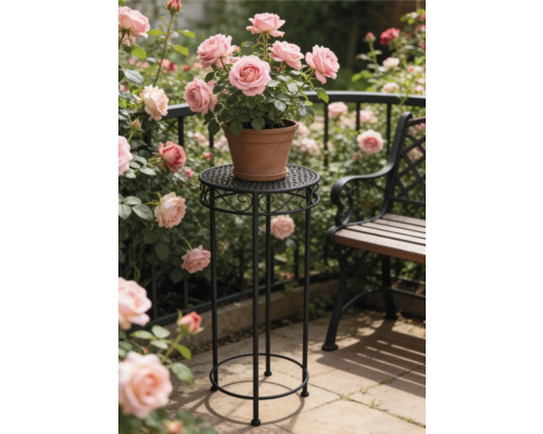 Porte-plante rond et haut en metal noir avec ornements portant un pot en terre cuite avec des roses roses dans un jardin lumineux pres du banc.