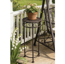 Hoher Blumenständer aus Metall mit Ornamenten auf einer Terrasse neben einer Gartenbank und einem Geländer.