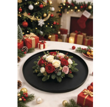 Composition florale de Noël avec roses, branches de pin et pommes de pin sur un plateau noir devant un sapin de Noël richement décoré.
