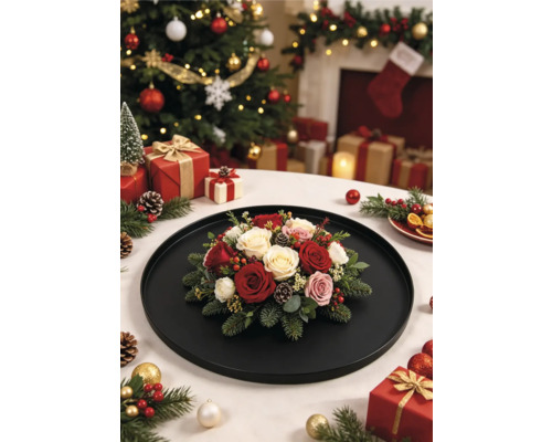 Composition florale de Noël avec roses, branches de pin et pommes de pin sur un plateau noir devant un sapin de Noël richement décoré.