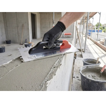 Hand mit Arbeitshandschuh trägt mit einer Zahnkelle Mörtel auf eine Betonwand auf einer Baustelle auf.