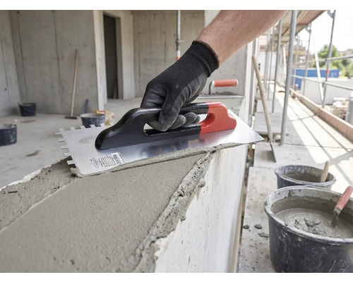 Hand mit Arbeitshandschuh trägt mit einer Zahnkelle Mörtel auf eine Betonwand auf einer Baustelle auf.