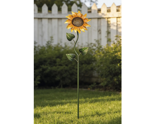 Metall-Gartenstecker in Sonnenblumenform in einem hellen Garten auf grünem Rasen vor Büschen und einem weißen Zaun.