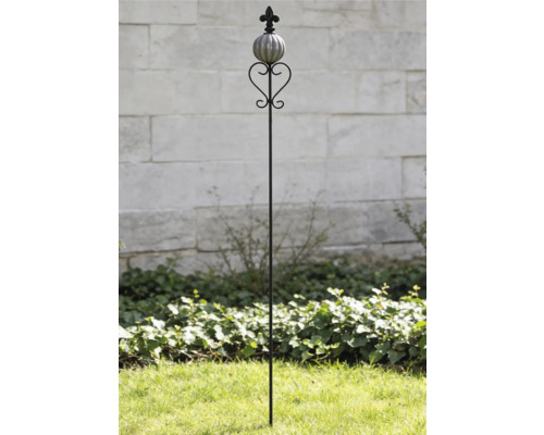 Tige de jardin décorative en métal avec boule en céramique, élément en forme de cœur et fleur de lys devant un mur en pierre.