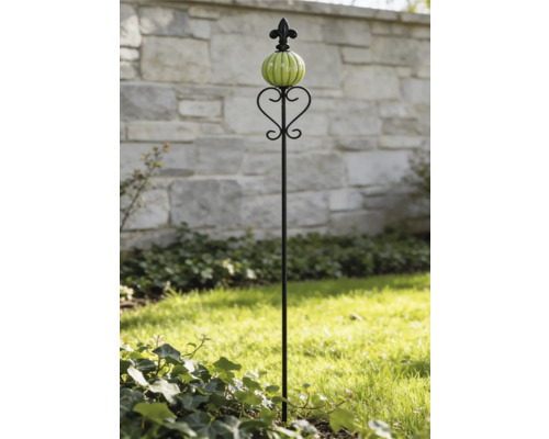 Schwarzer Gartenstab aus Metall mit grünem Glaskopf und Herzdekor vor einer Steinmauer.