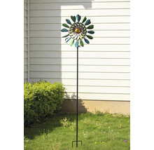 Éolienne de jardin en métal en forme de fleur sur une tige, placée dans un jardin devant un mur et un buisson.