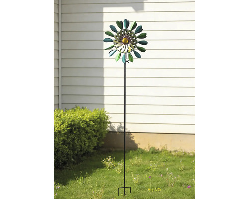 Éolienne de jardin en métal en forme de fleur sur une tige, placée dans un jardin devant un mur et un buisson.