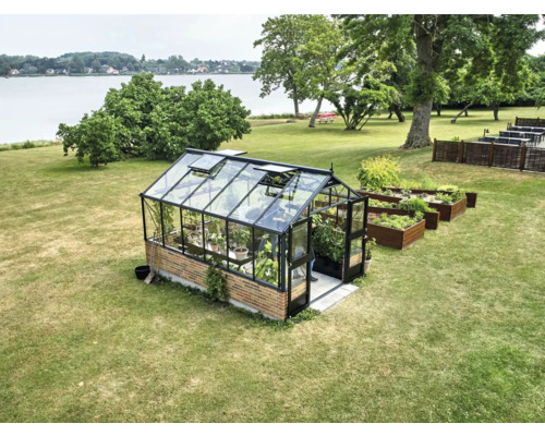 Gewächshaus mit schwarzem Aluminiumrahmen und Ziegelfundament auf Rasenfläche am See, neben hölzernen Hochbeeten.