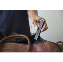 Une main tient un capteur mobile noir avec une dragonne au-dessus d'un sac en cuir marron.