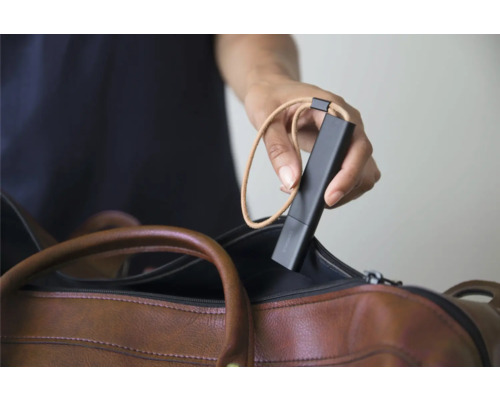 Une main tient un capteur mobile noir avec une dragonne au-dessus d'un sac en cuir marron.