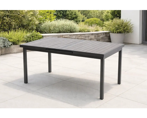 Table de jardin extensible moderne gris foncé sur une terrasse en pierre claire, entourée de plantes et d'un muret en pierre naturelle.