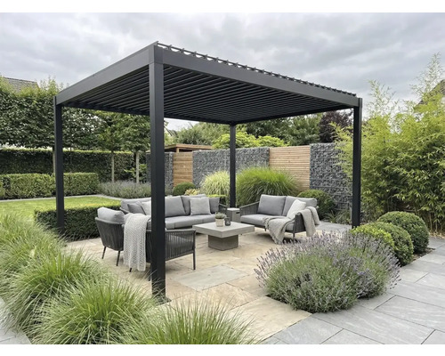 Pergola bioclimatique moderne en aluminium au-dessus d'un salon de jardin gris sur une terrasse en pierre, entourée de verdure.