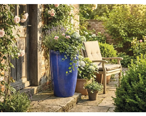 Grand pot de fleurs bleu en céramique sur terrasse en pierre, porte en bois, chaise de jardin et fleurs dans un jardin lumineux et soigné.