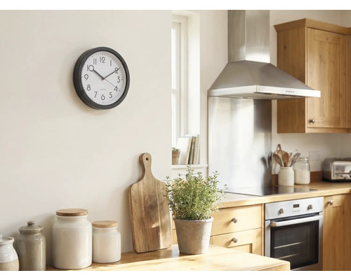 Helle Küche mit einer runden Wanduhr an der Wand, Holzschneidebrett, Kräutertopf und Aufbewahrungsgläsern auf der Arbeitsplatte.