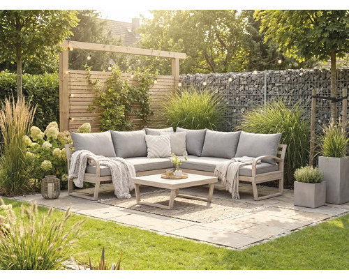 Eck-Gartenlounge aus Holz mit grauen Polstern, Holztisch und Teppich auf einer hellen Steinterrasse umgeben von Gräsern und Gabionenwand.