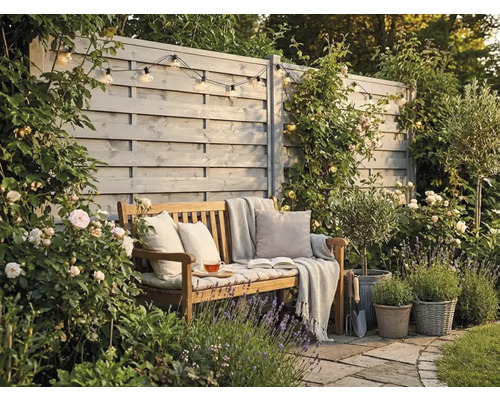 Coin de jardin accueillant avec banc en bois, coussins et couverture devant une clôture en bois grise, entouré de roses et de lavande.