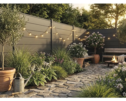 Jardin moderne avec clôture grise, pavés en pierre naturelle, plantes en pots en terre cuite, guirlande lumineuse et brasero allumé.