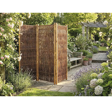 Paravent de jardin pliable à trois panneaux en osier dans un jardin fleuri avec terrasse en pierre et banc en bois.