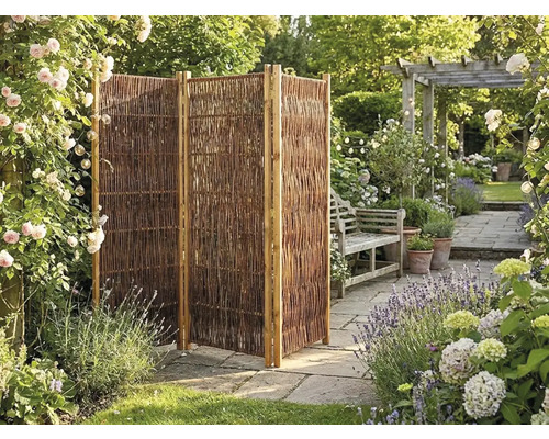 Paravent de jardin pliable à trois panneaux en osier dans un jardin fleuri avec terrasse en pierre et banc en bois.