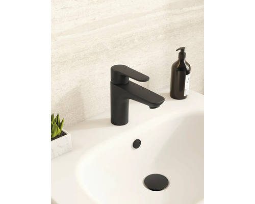 Mitigeur de lavabo moderne noir mat sur un lavabo blanc dans une salle de bains lumineuse avec distributeur de savon noir et petite plante.