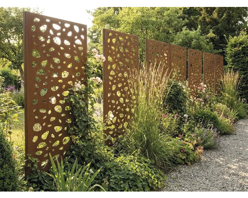 Sichtschutzwand aus Corten-Stahl mit organischem Lochmuster im Garten zwischen Stauden und Gräsern an einem Kiesweg.